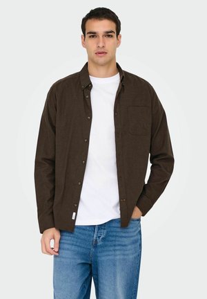 Camisa marrón de botones con un solo bolsillo en el pecho, llevada sobre una camiseta blanca. Combinada con jeans azules, presenta un cuello clásico y la tapeta de botones.
