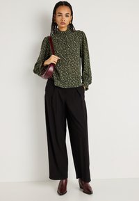 Πράσινη floral μπλούζα με υψηλή λαιμόκοψη και μακρύ μανίκι, συνδυασμένη με μαύρο παντελόνι με φαρδιές leg και μποτάκια σε μπορντώ τόνο με τακούνι; κρατάει μια ταιριαστή τσάντα ώμου.