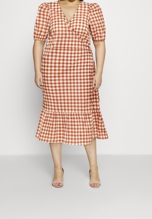 Mujer vistiendo un vestido cruzado de cuadros vichy en color óxido y blanco, con mangas abullonadas y volante en el dobladillo, combinado con tacones nude con pulsera en el tobillo.