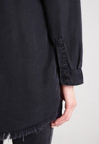 Camicia in denim scuro con orlo sfrangiato, polsini con bottoni e vestibilità rilassata, caratterizzata da una texture liscia e un design sobrio.