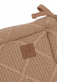 Geweven beige textiel met een diagonaal patroon, voorzien van een bruine leren label met een gestileerde "J" en een stoffen band.