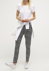 Žena stojí oblečená v bílém tričku, bílém svetru uvázaném v pase, štíhlých kalhotách s černobílým vzorem a bílých teniskách na světlém pozadí.