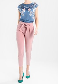 Bavlněná modrá blůza s květinovým potiskem a krátkými rukávy, doplněná o upnuté kalhoty v barvě broskve se zavazováním vpředu. Zelené lodičky na vysokém podpatku.