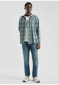 Zila kvadrātveida flaneļa krekls, uzvilkts virs balta grafiskā t-krekla, apvienots ar gaiši ziliem džinsiem un pelēkiem sporta apaviem, stāvot uz neitrāla fona.