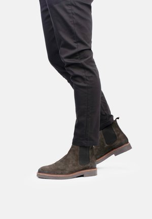Botas Chelsea de ante marrón con paneles laterales elásticos, con puntera redondeada y suela de color marrón claro contrastante, combinadas con pantalones negros.