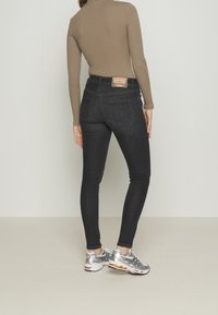 Jeans skinny de denim oscuro con cintura alta, bolsillos traseros y parche de marca. Combinados con una camiseta larga beige y zapatillas deportivas plateadas.