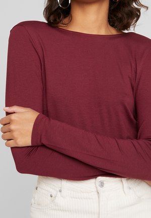 Burgunderfarbenes Langarmshirt aus weichem Baumwollmischgewebe mit rundem Ausschnitt und enganliegenden Ärmeln, kombiniert mit hellen Cordhosen.