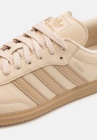 Beige läder Adidas Samba-sneakers med texturerad ovandel, tre tonala ränder och en gummisula med ett sicksackmönster.