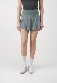 A teal athletic skort with a wide, ribbed waistband and a flared silhouette, paired with white athletic shoes and crew socks.