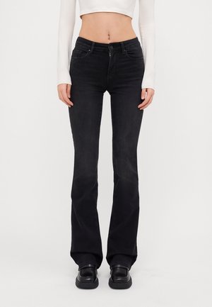 Kobieta ubrana w czarne dżinsy typu bootcut z wysokim stanem, biały crop top z długim rękawem oraz czarne masywne buty, stojąca na jednolitym tle.