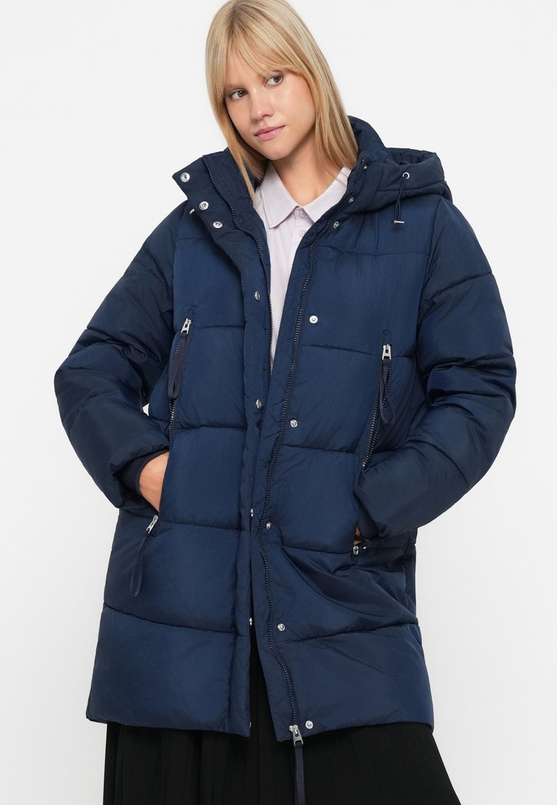 Cappotto imbottito blu navy con cappuccio, tasche con zip, chiusura frontale con zip e bottoni automatici, e una texture trapuntata. Presenta una vestibilità ampia e polsini elasticizzati.