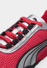 Raudonas ir sidabro spalvos sportbačiai su juodu logotipu, raudonais raišteliais, įvertais per juodus raištelių laikiklius, ir viršutine dalimi iš tinklelio audinio.