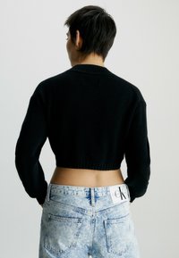 Schwarzer, kürzer geschnittener Pullover mit geripptem Saum und langen Ärmeln, kombiniert mit hellblauen Denim-Jeans, die ein Logopatch auf der Rückseite haben.