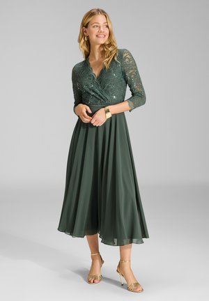 Femme portant un haut en dentelle vert foncé et une robe à jupe fluide avec un bracelet doré et des sandales à talons hauts, debout devant un fond gris.