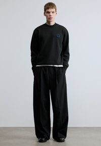Μαύρο φούτερ με υφασματικό στρογγυλό έμβλημα, συνδυασμένο με φαρδιά μαύρα παντελόνια και μαύρα παπούτσια. Λείο ύφασμα, χαλαρή σιλουέτα.