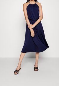 Kvinna står i en ärmlös marinblå halterneckklänning med knytbar midja, iförd svarta klacksandaler mot en enkel vit bakgrund.