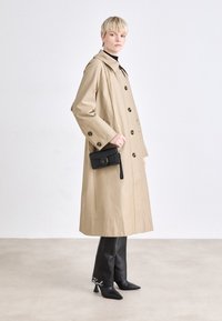 Trench beige con grandes botones negros, usado sobre pantalones negros ajustados y tacones de punta. Pequeño bolso negro con cadena dorada.