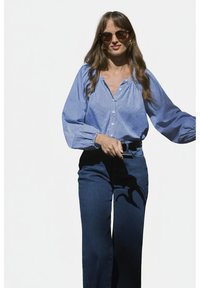 Blauw-wit gestreepte blouse met pofmouwen, gecombineerd met donkerblauwe wijde jeans. Model draagt een zonnebril en een riem.