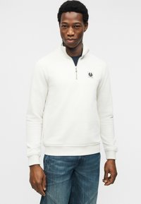 Άνδρας που φοράει λευκή μπλούζα με φερμουάρ έως τη μέση και λογότυπο στο στήθος, με μπλε τζιν, στέκεται μπροστά σε απλό λευκό φόντο.