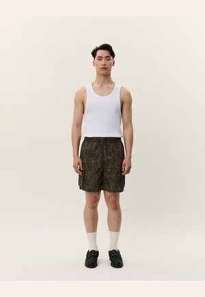 Un tânăr stă cu fața spre înainte, purtând un tricou fără mâneci alb, pantaloni scurți cu un model închis, șosete albe și pantofi negri, pe un fundal simplu.