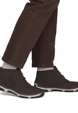 Personne portant un pantalon marron foncé et des chaussures montantes marron foncé à lacets avec semelles blanches et chaussettes de couleur claire.