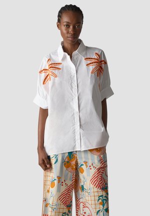 Woman wearing a white button-up shirt with orange leaf designs on sleeves and colorful patterned wide-leg pants against a plain background.