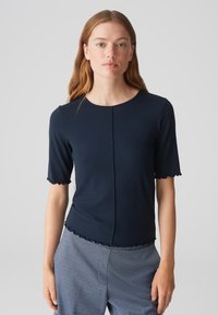 Marineblauwe, kortgebouwde top van een gestructureerde stof, met golvende randen langs de mouwen en de zoom, en een centrale stiknaad details.