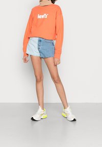 Persona vistiendo un sudadera naranja de Levi's, pantalones cortos de mezclilla bicolor azul y zapatillas blancas con acentos en negro y amarillo neón.