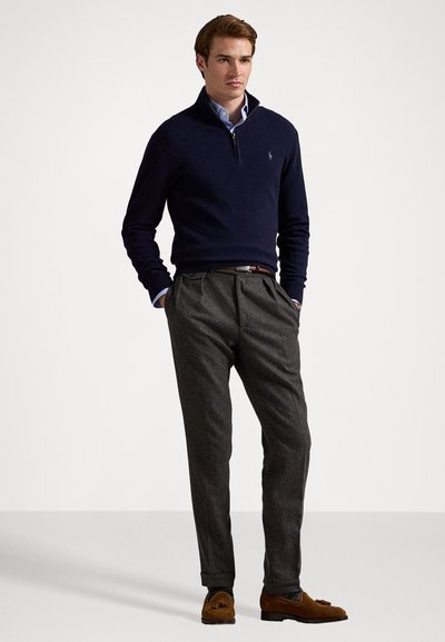 Marineblauwe pullover trui met ritssluiting bij de kraag, gedragen over een lichtblauw overhemd met kraag, gecombineerd met grijze geplooide broeken en bruine suède loafers.
