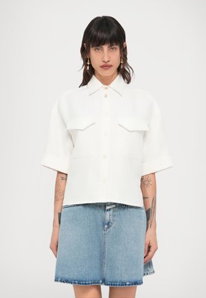 Chemise blanche à manches courtes avec col, deux poches poitrine et ouverture boutonnée à l'avant ; confectionnée dans un tissu léger, associée à une jupe en denim bleu.