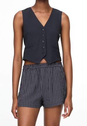 Ærmeløs sort knapvest parret med sort-hvide vertikalt striped shorts på en stående person.