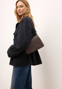 Brązowa skórzana torebka o gładkiej teksturze, prostokątnym kształcie i złotych akcentach, noszona na ciemnej kurtce i niebieskich dżinsach.