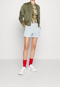 Olivgrön bomberjacka, batikfärgad brun topp, ljusblå shorts, röda strumpor och vita chunky sneakers. Släta material och avslappnad passform.