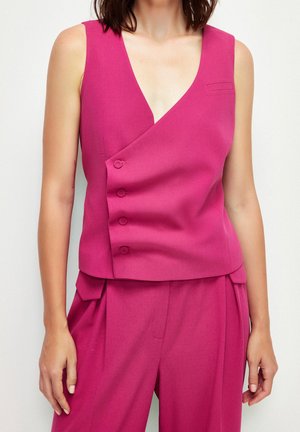 Femme portant un gilet magenta sans manches avec une fermeture asymétrique à boutons et un pantalon plissé taille haute assorti sur un fond uni.