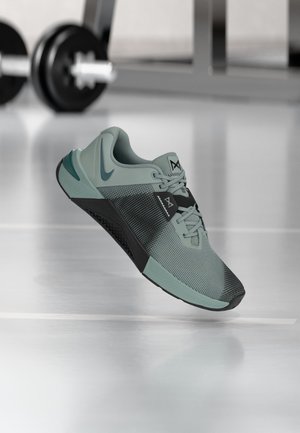 Chaussure de sport turquoise clair et noire suspendue en l'air dans une salle de sport, avec des haltères et des poids flous en arrière-plan sur un sol brillant.