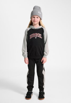 Kind steht mit grauer Mütze, schwarz-grauem Hoodie mit dem Schriftzug „HUMMEL“, schwarz-grauen Hosen und schwarzen Turnschuhen vor weißem Hintergrund.