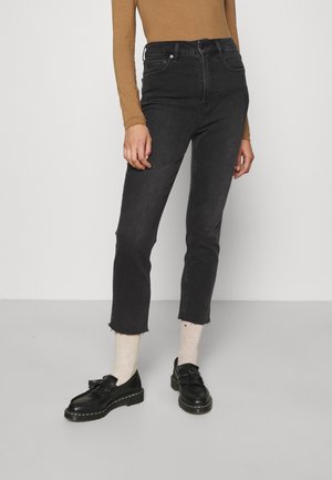 Kobieta w czarnych, krótkich spodniach z wysokim stanem, beżowej bluzce z długim rękawem, kremowych skarpetach i czarnych mokasynach z frędzlami stoi na szarej podłodze.