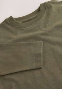 Bluză cu guler rotund verde măsliniu realizată din material moale, cu guler ribanat și suprafață ușor texturată, cu cusături cusute.