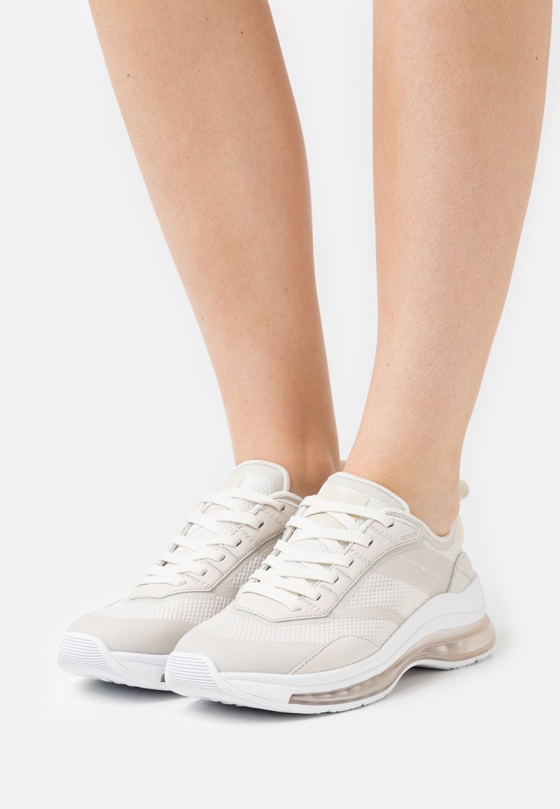 Jambes portant des baskets basses blanches avec des détails en mesh et en cuir, des lacets blancs et des semelles transparentes rembourrées sur fond blanc.