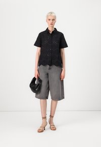 Camicia nera a occhio di pernice con maniche corte, orlo scallop, abbinata a culotte di denim grigio. Accessoriata con una borsa nera e sandali beige con lacci.
