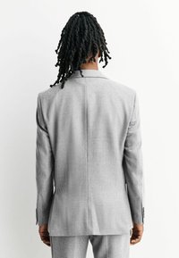 Person with long twisted hair wearing a light gray checked suit, standing with their back to the camera against a white background.