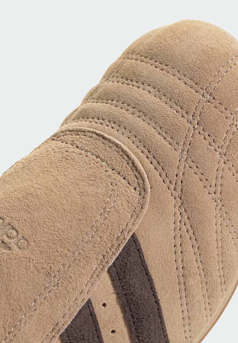 Zapato de ante beige con parte superior texturizada, presenta rayas de acento en marrón, diseño acolchado y área de tobillo acolchada para soporte.
