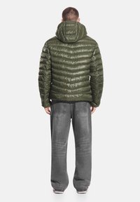 Oliwkowa kurtka puchowa z poziomym przeszyciem i kapturem, zestawiona z luźnymi szarymi dżinsami. Na kurtce widoczna jest gładka, błyszcząca faktura.