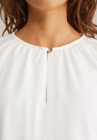 Weiße Bluse mit rundem Ausschnitt, die ein gerafftes Stoffdesign aufweist. Ein goldener Knopfverschluss am Hals stellt ein auffälliges Detail dar.