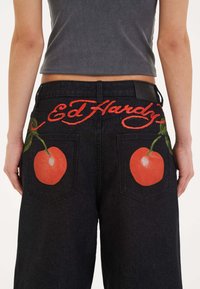Jeans de mezclilla negra con texto bordado en rojo "Ed Hardy" encima de dos grandes diseños de frutas naranja en los bolsillos traseros.
