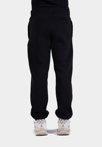 Pantaloni de trening negri cu o croială lejeră, fabricați dintr-un material moale. Prezintă un elastic la talie și buzunare laterale. Au fost asortați cu adidași albi.