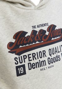 Hoodie gris clair avec un imprimé graphique en bleu foncé et marron. Le texte indique "Jack & Jones Superior Quality Denim Goods" avec un élément décoratif en forme de ruban.