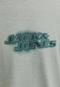 Texte vert « JACK & JONES » avec un effet d'ombre imprimé sur un fond en tissu gris clair, probablement une partie d'un vêtement.