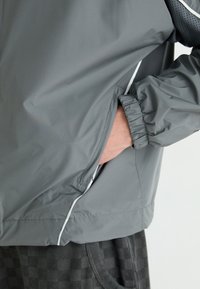 Veste coupe-vent légère grise avec des poignets élastiques, des poches latérales et des accents blancs réfléchissants le long des coutures. La texture semble lisse et durable.