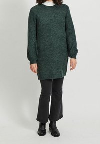 Abito maglione verde scuro a maglia con lunghe maniche a sbuffo, polsini a costine e orlo dritto, abbinato a pantaloni neri a palazzo e stivali alla caviglia.